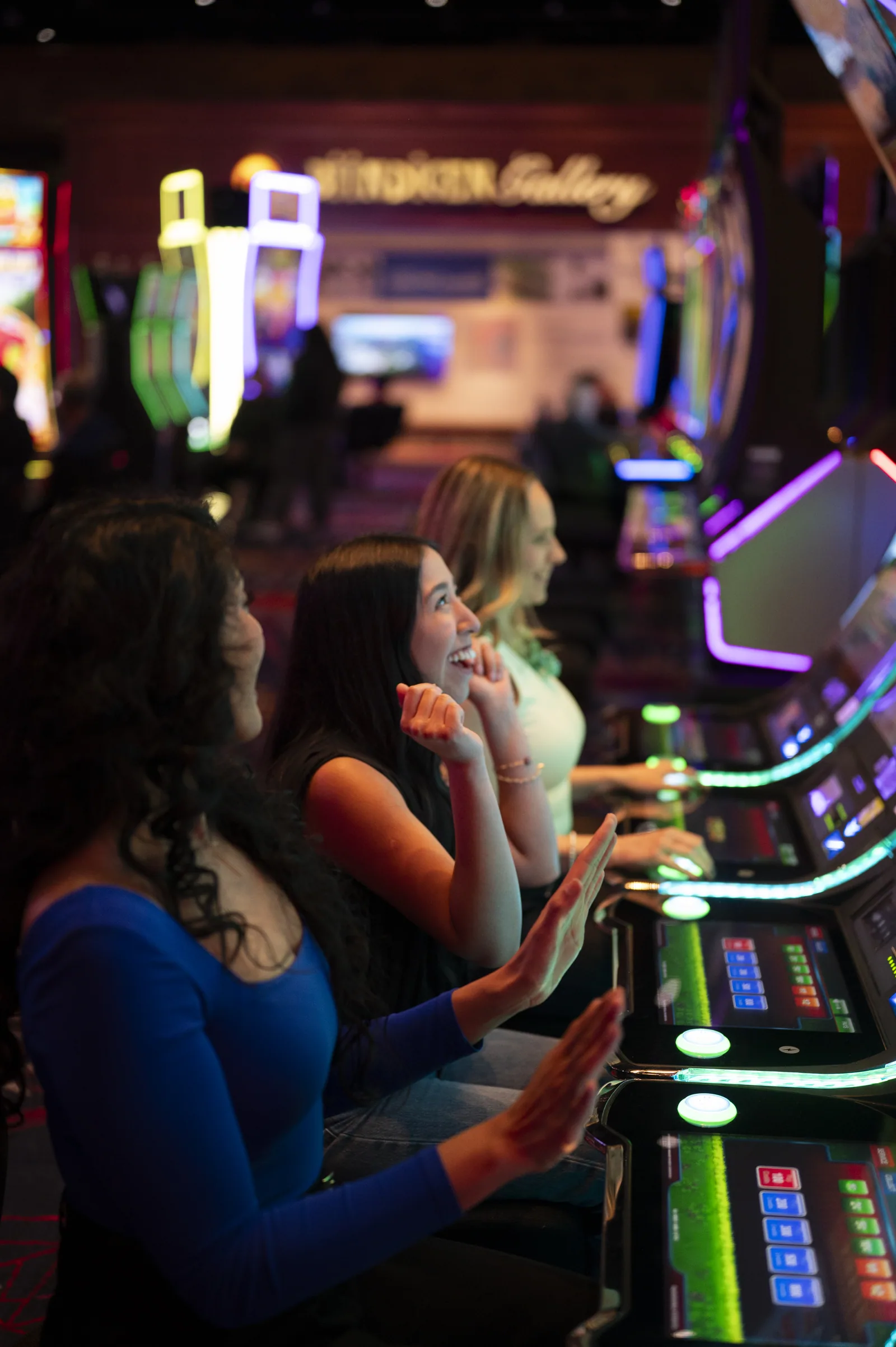 Three women laugh and clap while playing modern slot machines in a casino