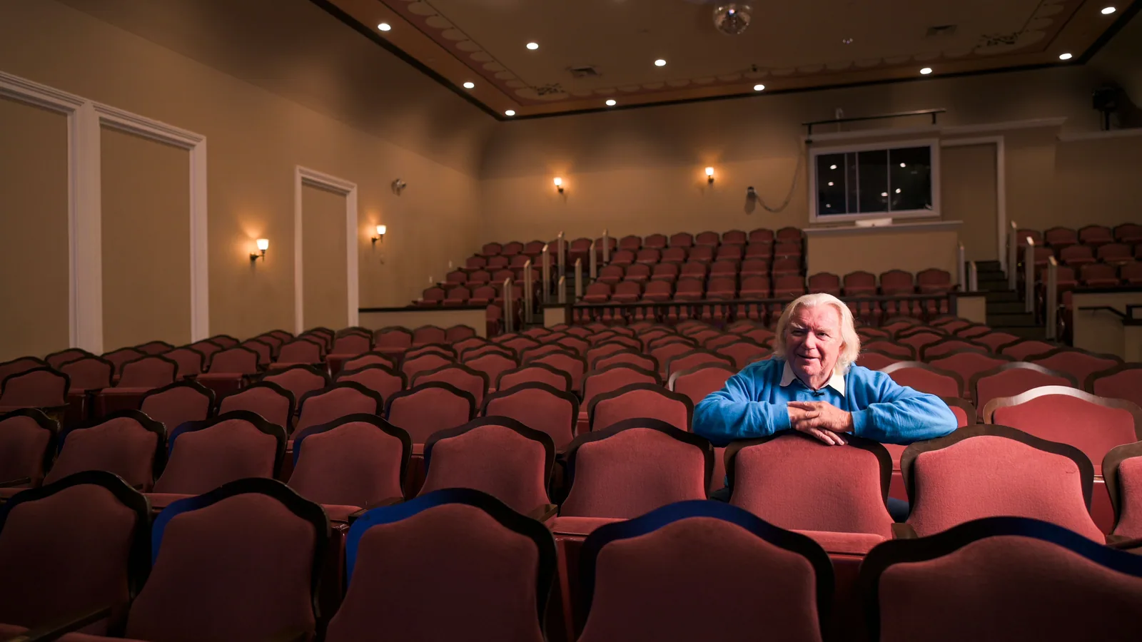 Man with white hair in a blue shirt sits in a red velvet seat in a theater