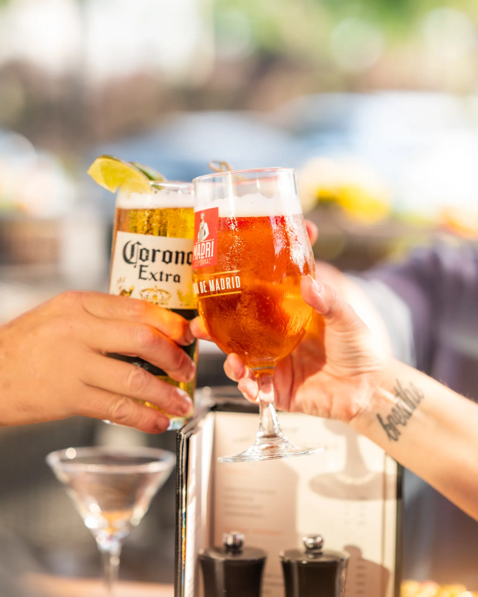 Two people clinking glasses of beer, one Corona, one Madri, on a sunny patio