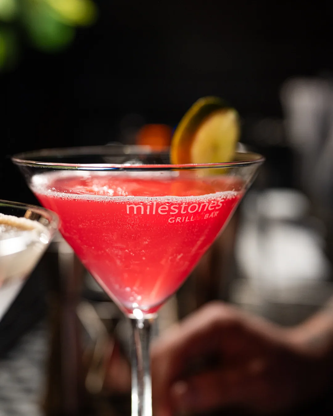 Milestones martini glass filled with a pink cocktail, lime garnish, bar setting