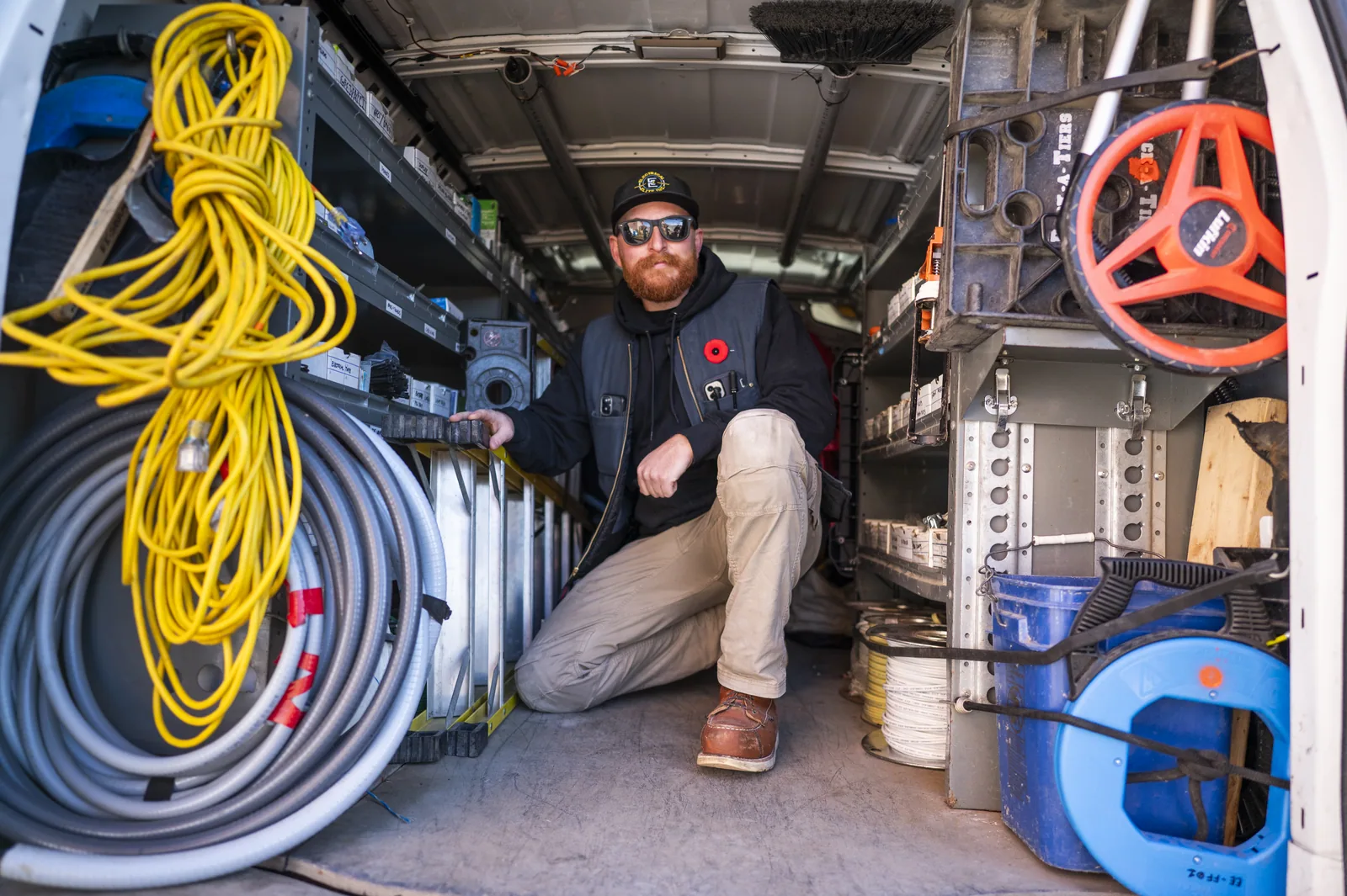 Male electrician with beard, sunglasses, and work vest kneeling inside a packed service van