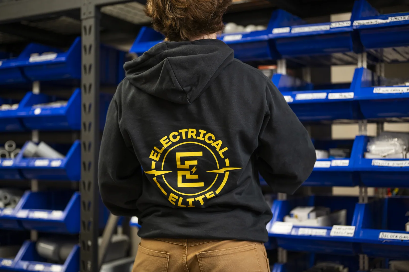 Person from behind wearing Electrical Elite hoodie in front of parts storage