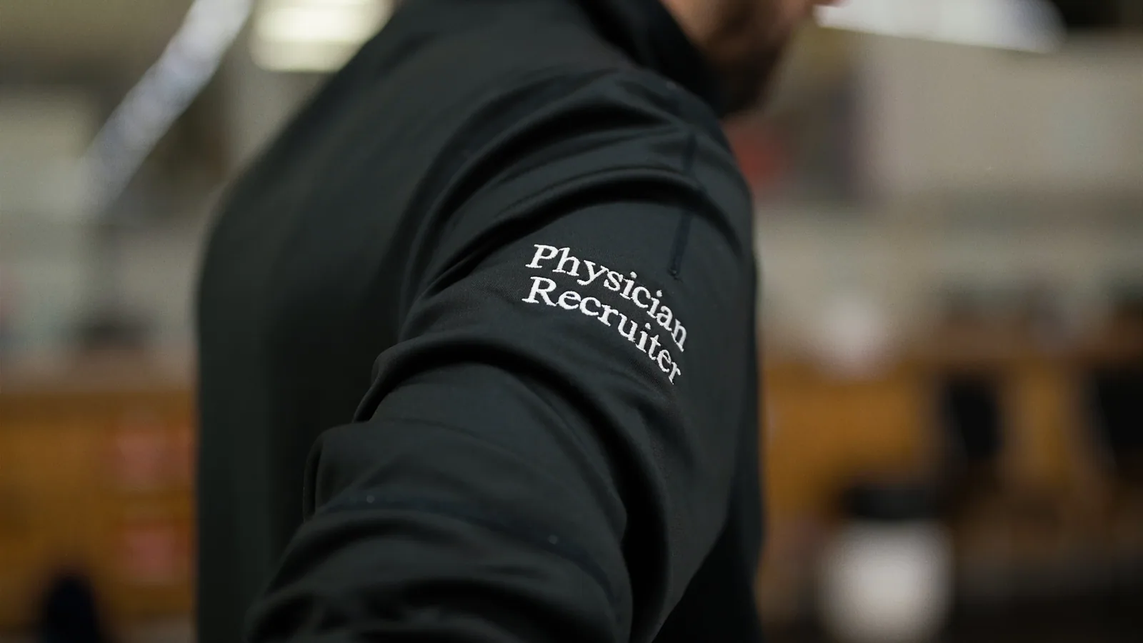 Person's arm in a black jacket with 'Physician Recruiter' embroidered on the sleeve