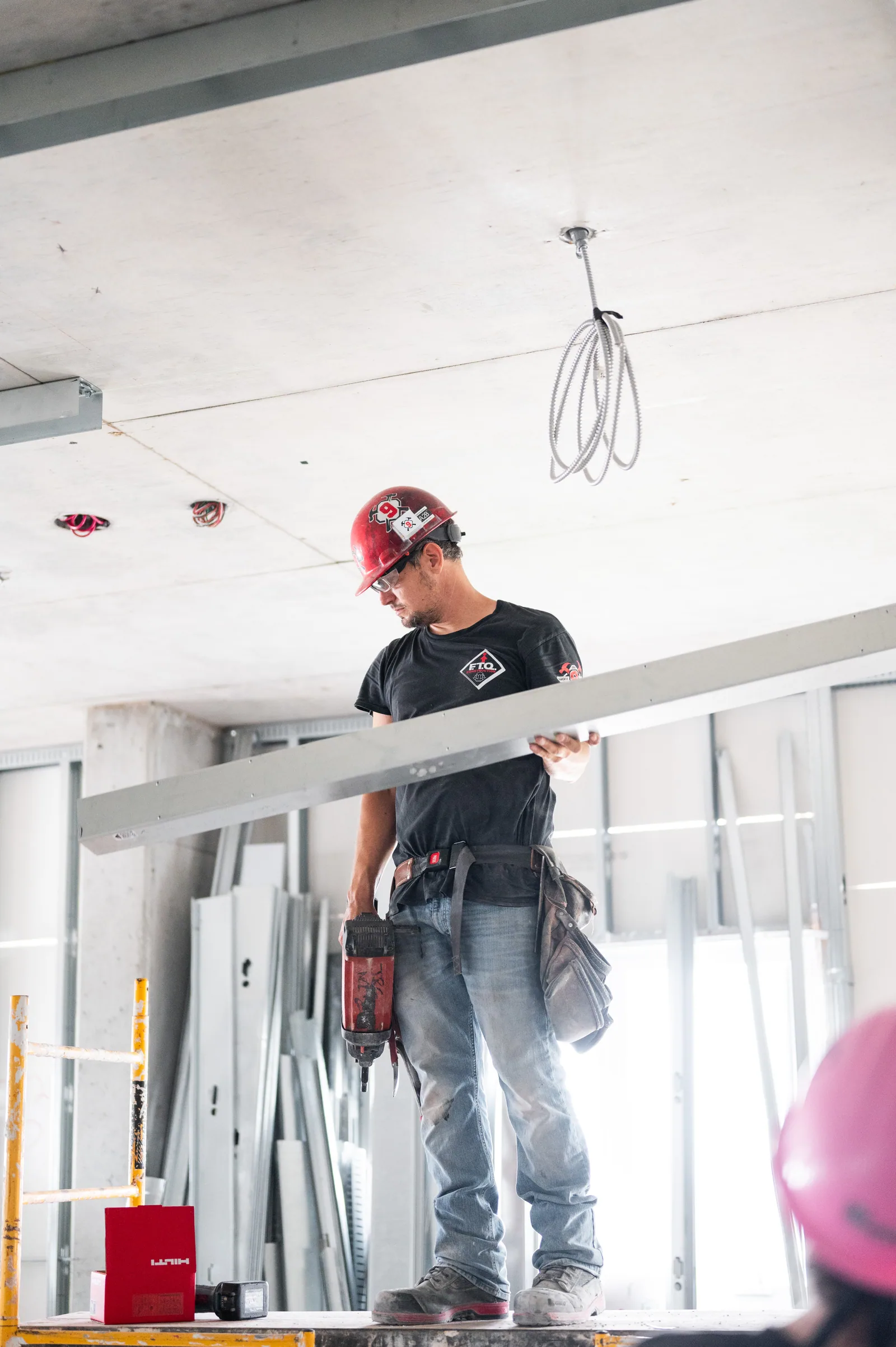 Construction worker in hard hat holds metal framing piece and power tool on scaffolding