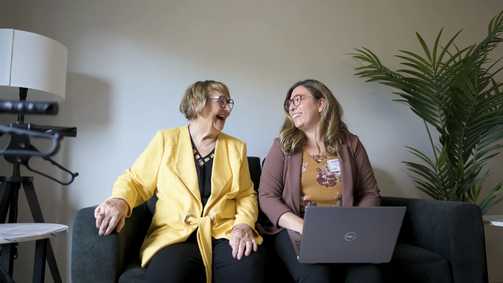 Two smiling women, one older, one younger, laughing on a couch with laptop