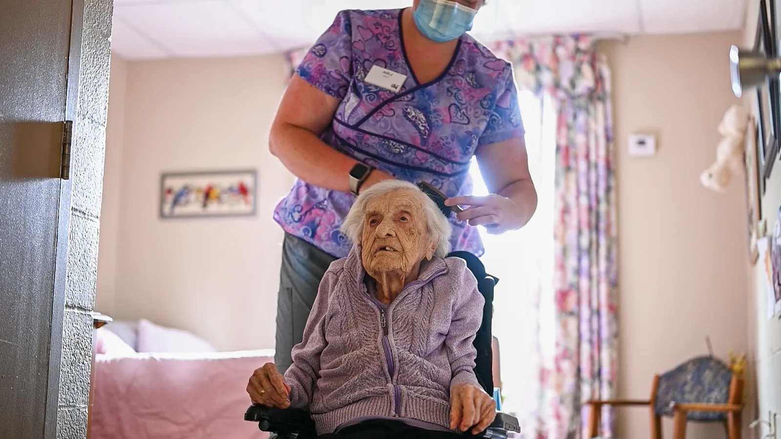 Healthcare professional in scrubs brushing an elderly woman's hair in a care home